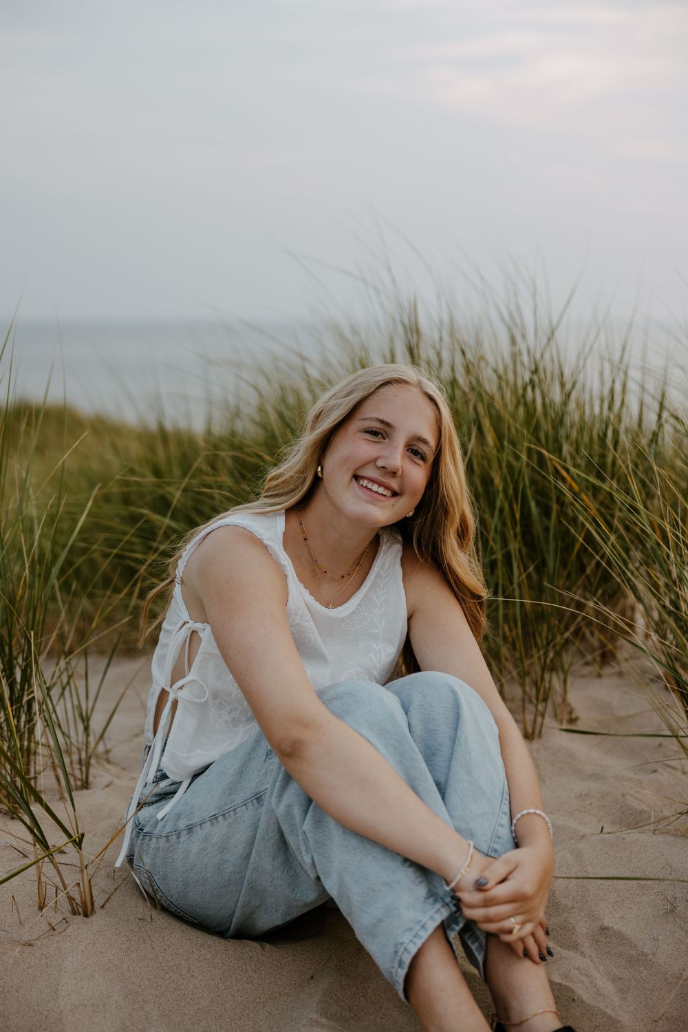 A young woman with long blonde hair smiles while sitting on a sandy beach surrounded by tall grass. Fort Worth Texas senior photographer.