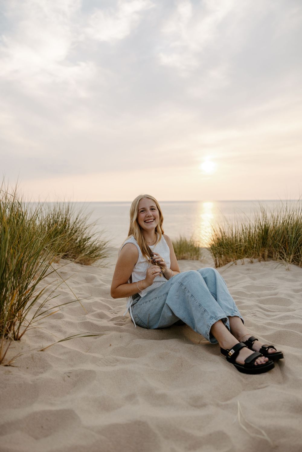 A young woman sits on a sandy beach, surrounded by tall grass, with the sun setting in the distance. Fort Worth Texas senior photographer.