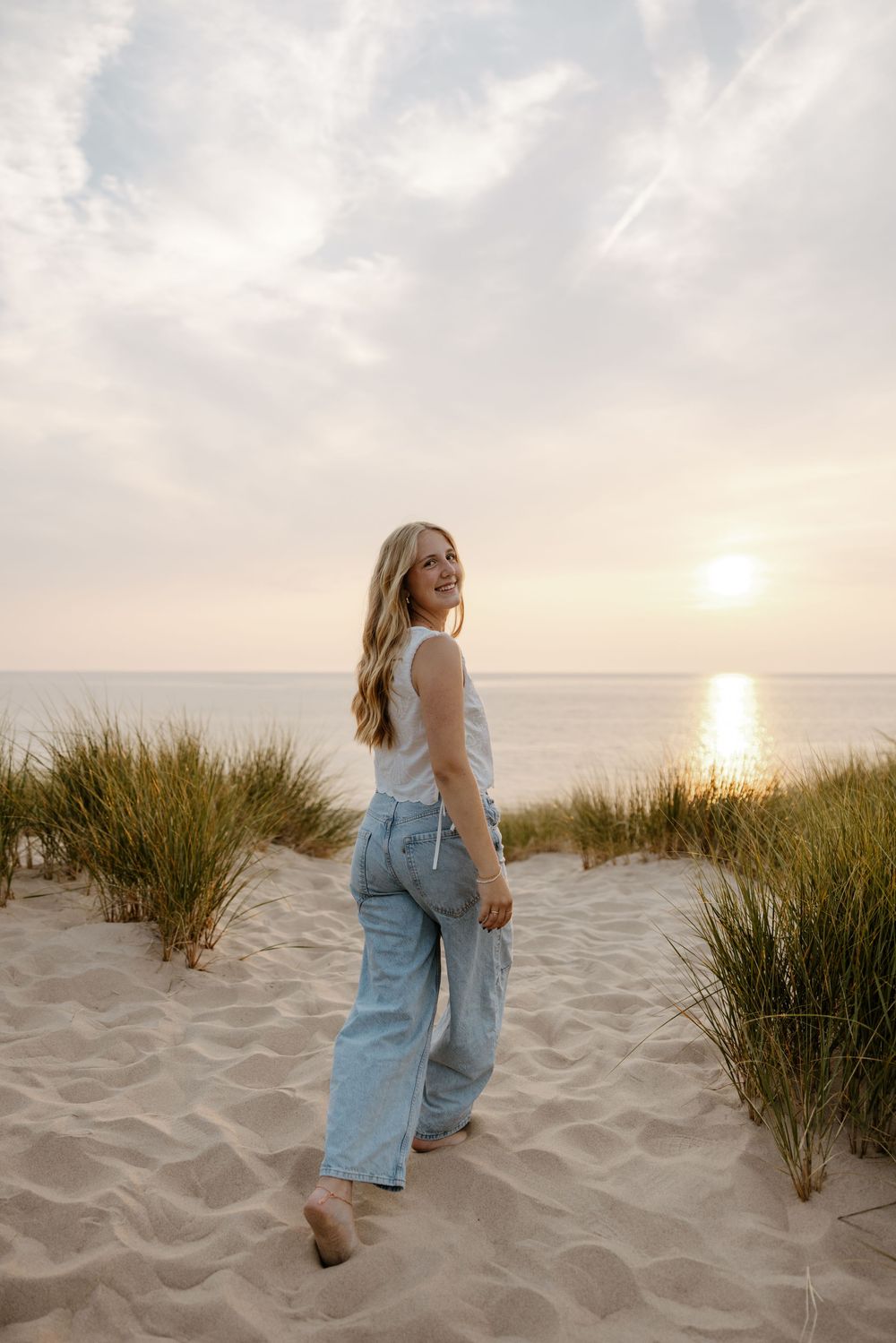 A woman with long blonde hair walks barefoot on a sandy beach at sunset, with tall grass and a calm sea in the background. Fort Worth Texas