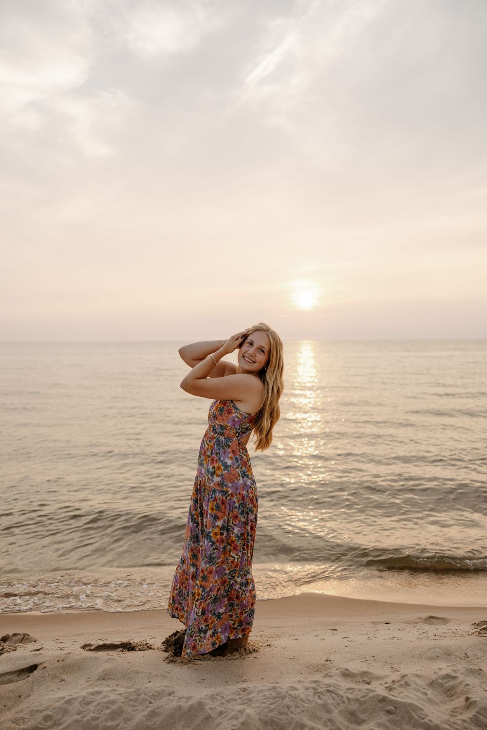 A woman in a colorful floral dress stands on a beach, gazing towards the setting sun over the ocean. Fort Worth Texas senior photographer.