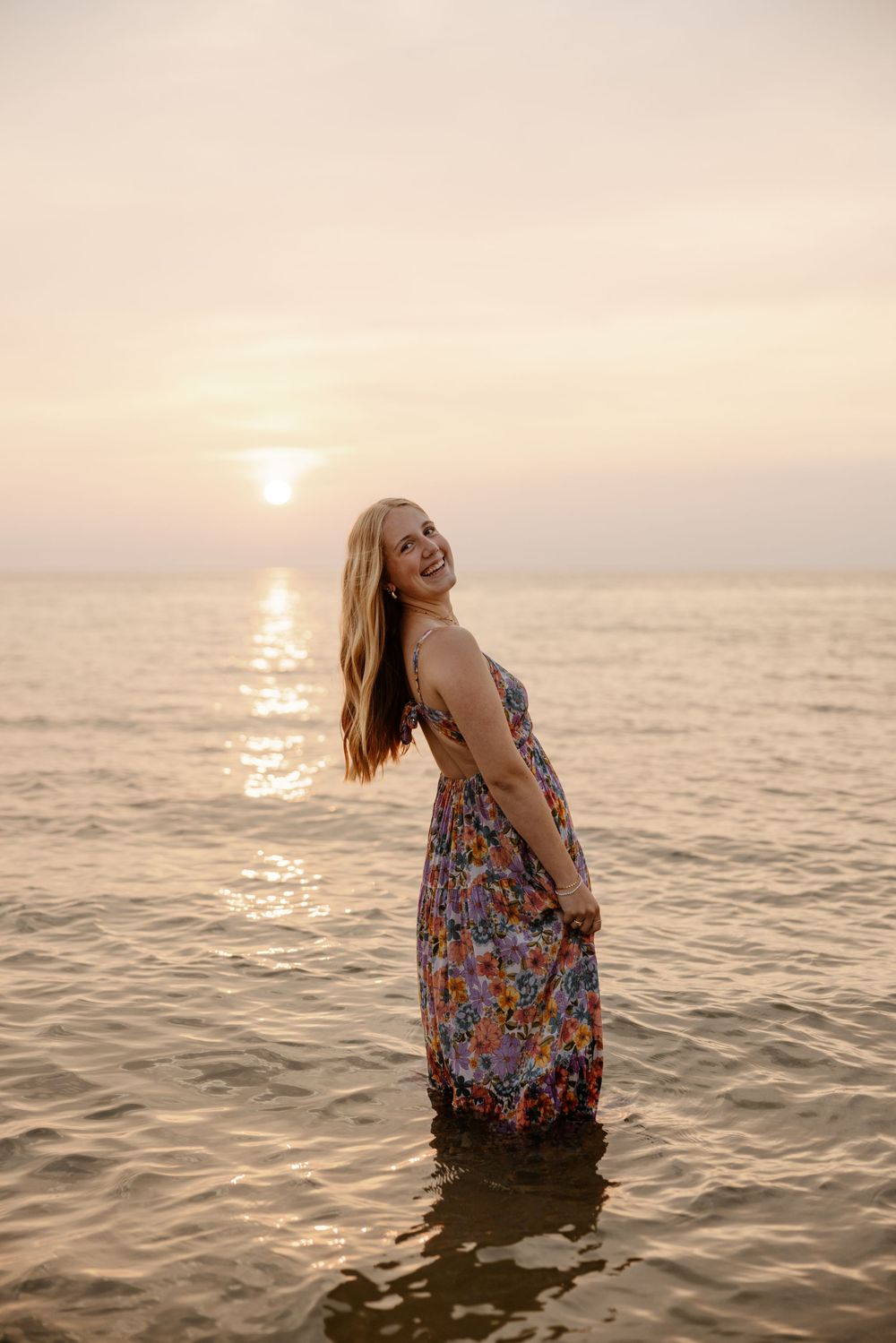 A woman in a colorful floral dress stands in the shallow water at sunset, with the sun's reflection on the water behind her. Fort Worth TX