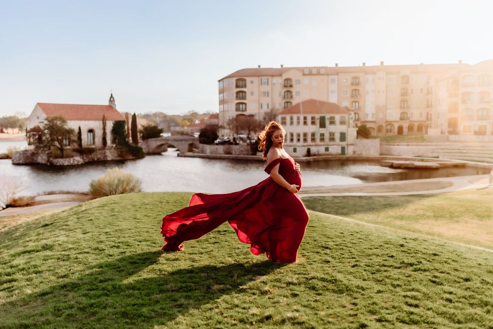 A flowing red dress billows in the wind on a grassy hill overlooking a coastal town at sunset.