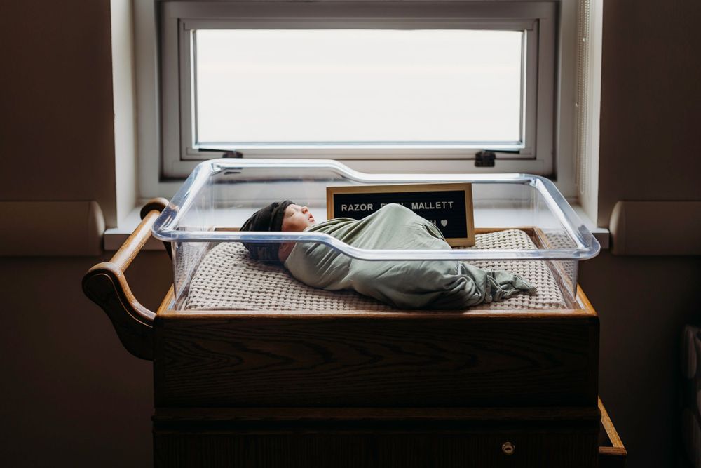 Newborn baby resting in hospital bassinet by window with soft natural light.