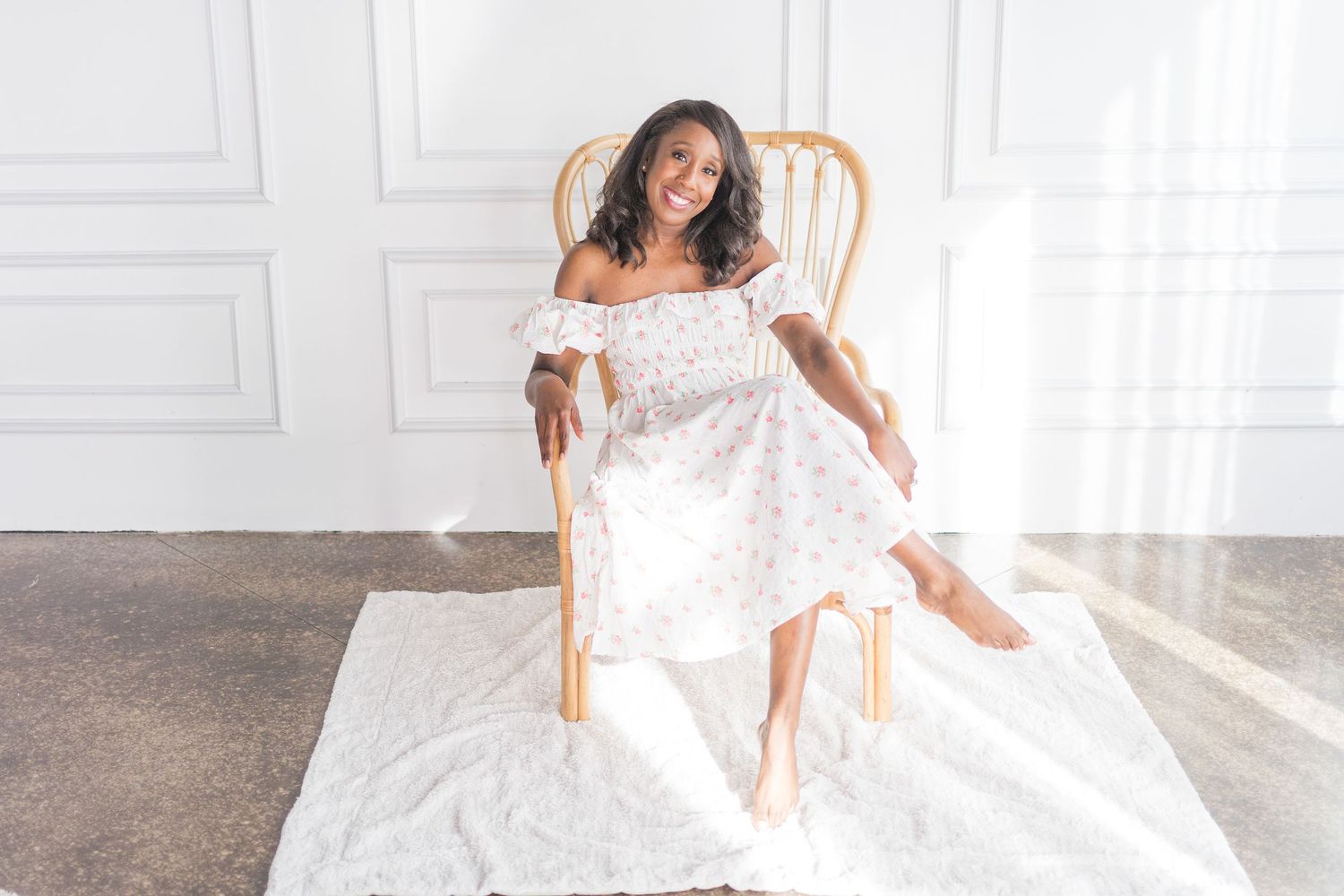 Someone in a white off-shoulder dress poses gracefully on a wooden chair against a bright white wall interior.