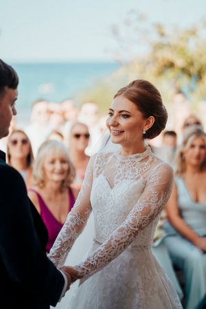 A bride smiles joyfully during her outdoor wedding ceremony wearing an elegant long-sleeved lace gown.
