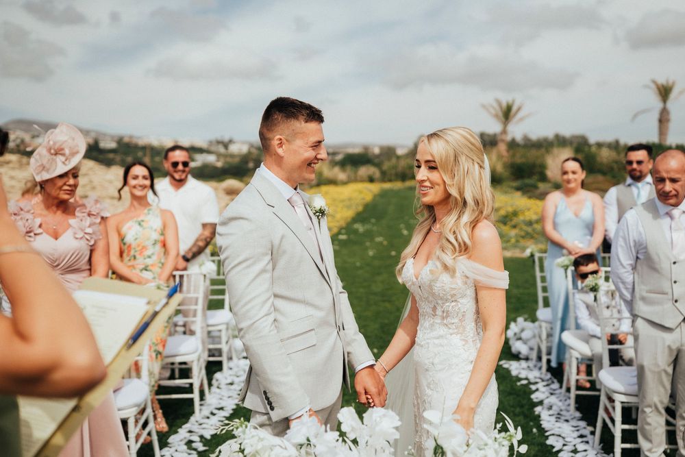 A joyful outdoor wedding ceremony in a lush field with palm trees and white chairs under cloudy skies.