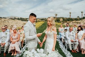 A couple exchanges vows during an outdoor desert wedding ceremony with guests seated in white chairs.