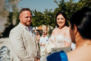 A wedding celebration outdoors with guests seated in white chairs and floral arrangements on a sunny day.
