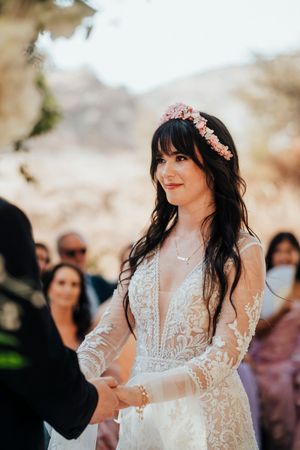 Beautiful bride in lace wedding dress with long sleeves and floral crown smiles during outdoor ceremony.