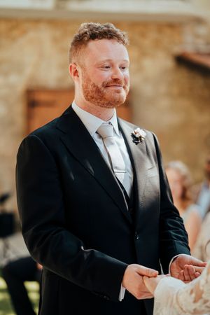 Wedding ceremony participant in formal black suit and white tie smiling during a special moment.