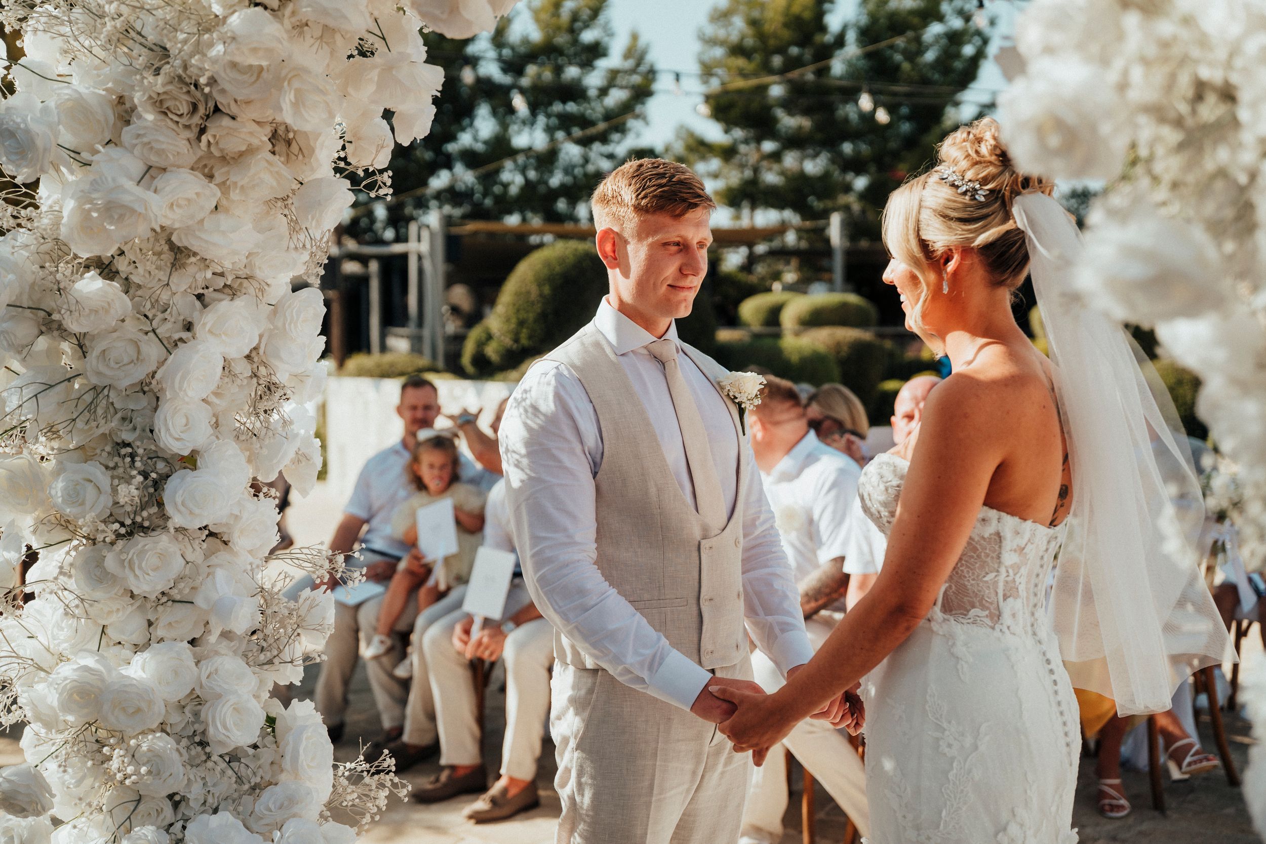 Couple holds hands during outdoor summer wedding ceremony under beautiful white floral arch.