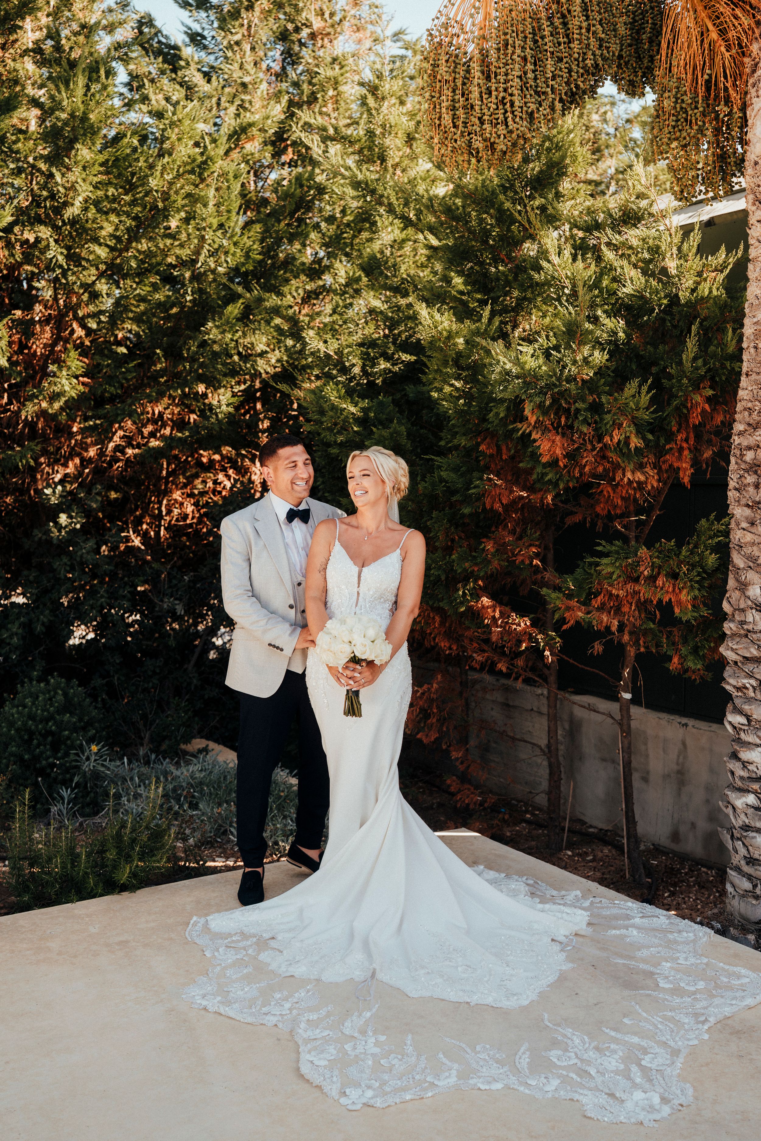 A couple poses together in formal wedding attire against a lush garden backdrop with palm trees and autumn foliage.