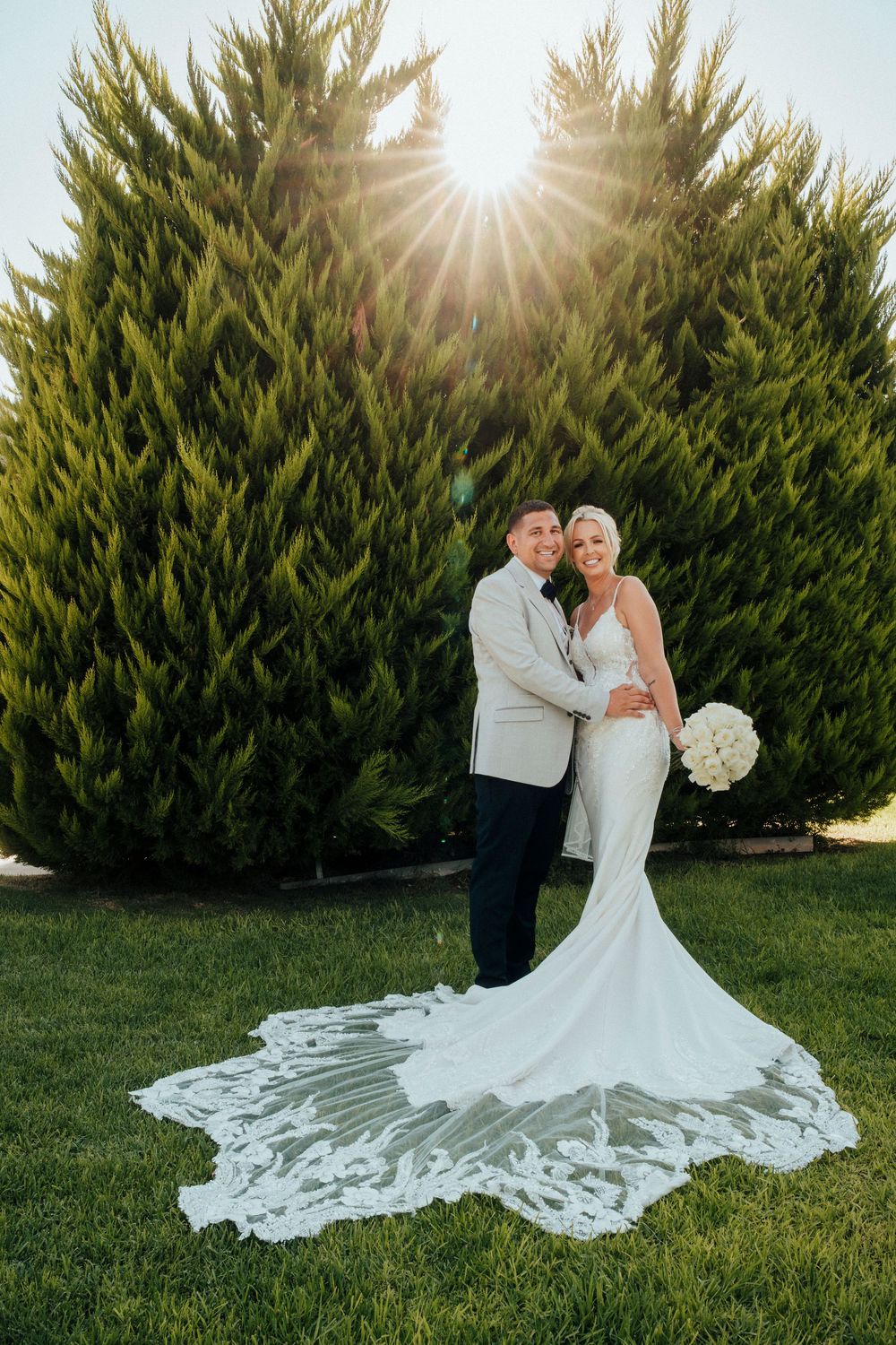 Wedding couple poses on green lawn with dramatic white lace train and sunburst through evergreen trees in background.