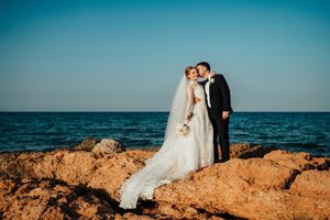 A wedding couple stands on rocky coastal cliffs overlooking turquoise ocean waters against a vibrant blue sky.