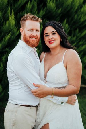 A couple in white attire embraces outdoors against a backdrop of green cypress trees.