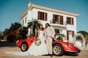 A couple poses with a vintage red Ferrari sports car in front of a Mediterranean-style white villa on a sunny day.