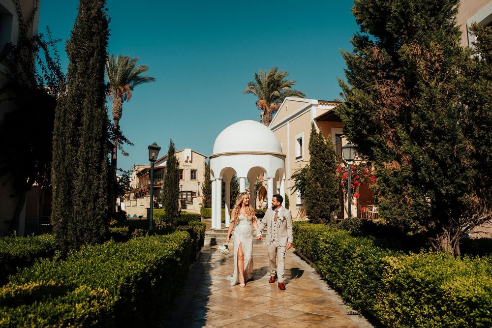 A couple walks along a Mediterranean-style garden path with palm trees and a domed building in the background.