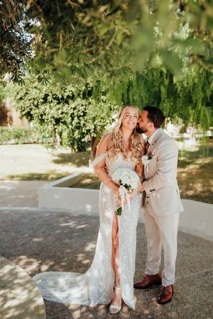 A couple shares intimate moments in a garden setting wearing wedding attire under dappled sunlight.