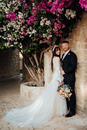 A wedding couple poses beneath a blooming bougainvillea tree against a stone wall at a Mediterranean venue.