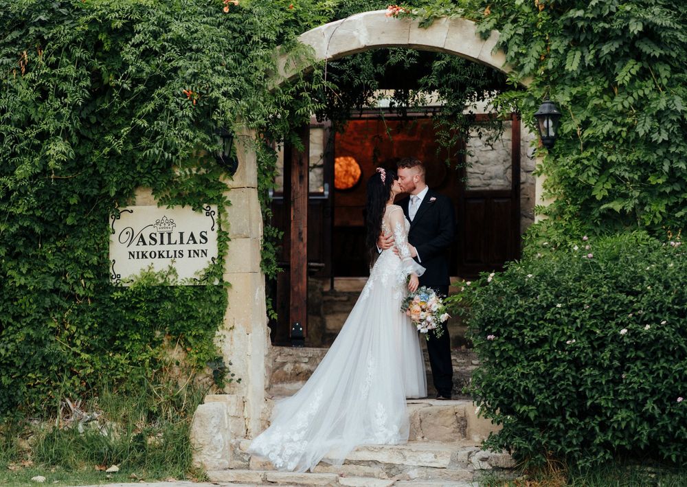 A bride in a flowing white gown poses in a rustic garden setting with stone walls and lush greenery.