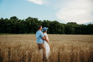 A couple embraces in a golden wheat field on a sunny summer day with trees in the background.