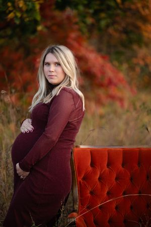 A person in a burgundy sweater poses on a red velvet couch outdoors with autumn foliage in the background.