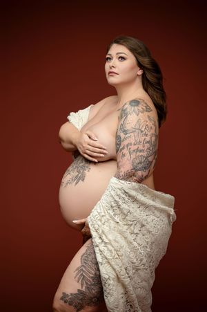 A maternity photoshoot features a cream lace dress and visible arm tattoos against a deep burgundy background.