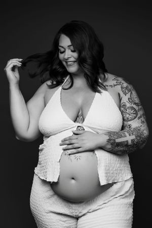 Black and white maternity photos showing a plus size woman in white lingerie displaying her pregnant belly with tattoos.