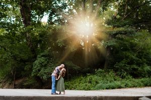 A couple walks along a sunlit path in a lush green forest with sunbeams streaming through the trees.