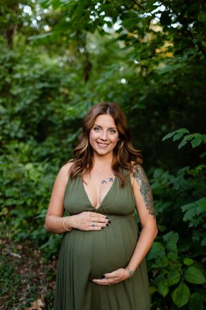 A pregnant woman in an olive green dress stands in a lush garden setting while gently cradling her baby bump.