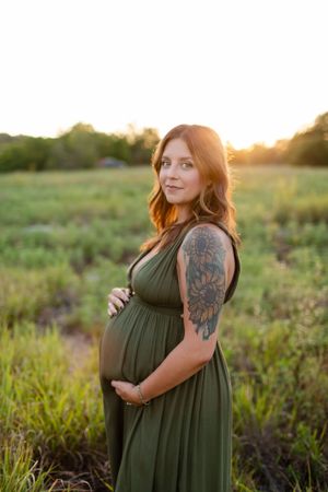 Expectant mother in olive green dress poses in golden sunlit field during sunset maternity photoshoot.