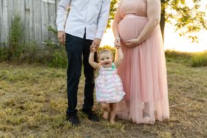 A small child in a pink dress stands between two adults during a sunset maternity photo session outdoors.