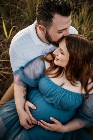 Intimate maternity photo of couple embracing in tall grass during golden hour, wearing white and blue outfits.