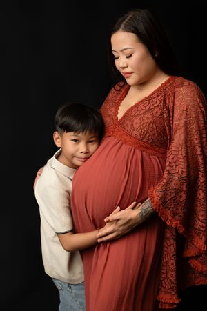 A maternity photo series showing a child in white embracing a pregnant figure wearing a rust-colored lace dress against black.