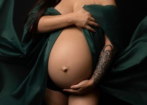Pregnant belly cradled by hands with intricate tattoo sleeve against dark emerald green fabric backdrop.