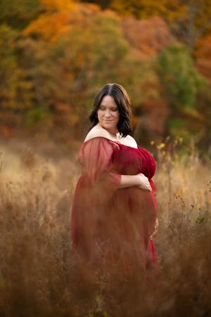 A pair in matching red dresses stand together in a field with fall colors in the background.