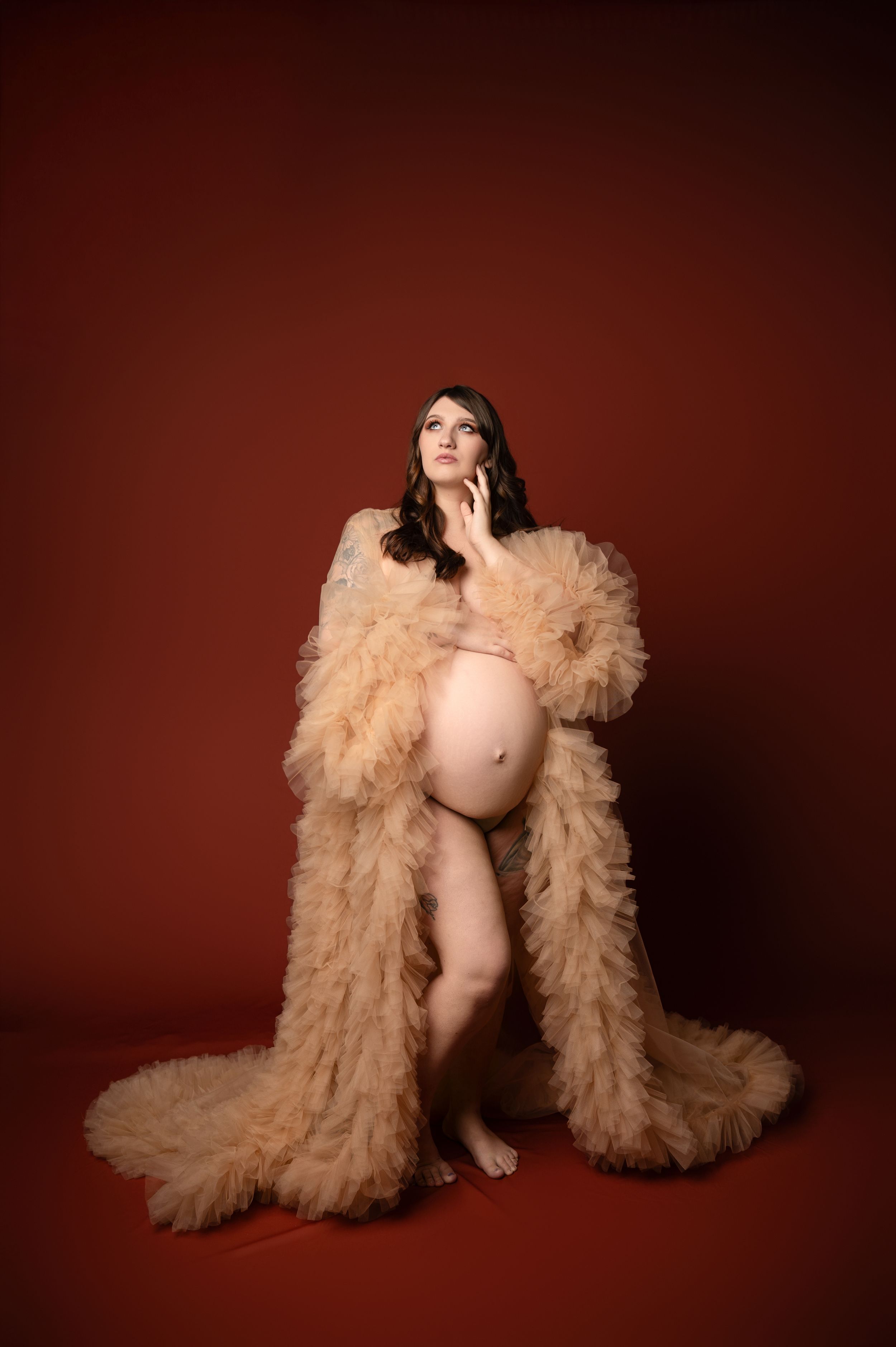 Artistic maternity photo featuring an elegant nude tulle robe in champagne color against a deep burgundy backdrop.