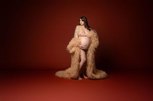 A pregnant figure poses in a beige fur robe against a deep red studio backdrop.
