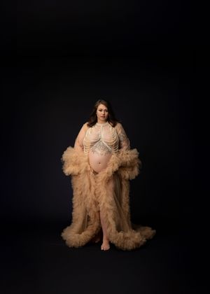 A figure in a romantic beige tulle robe poses against a dark background in an artistic maternity photoshoot.