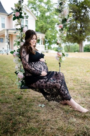 Expectant mother in black lace maternity dress sits on floral swing outdoors in garden setting.
