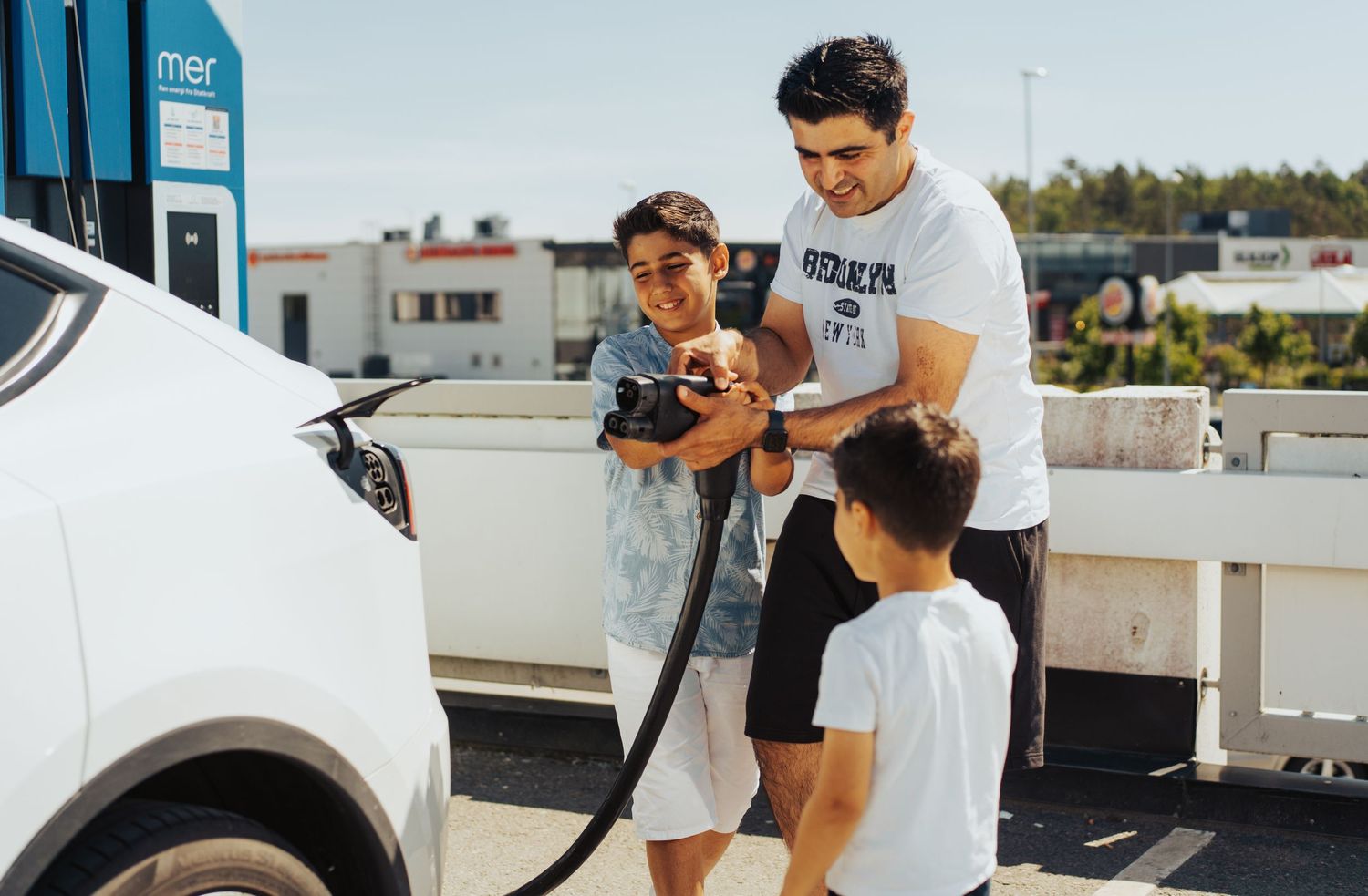 People gathered around a white car in a driveway on a sunny day.