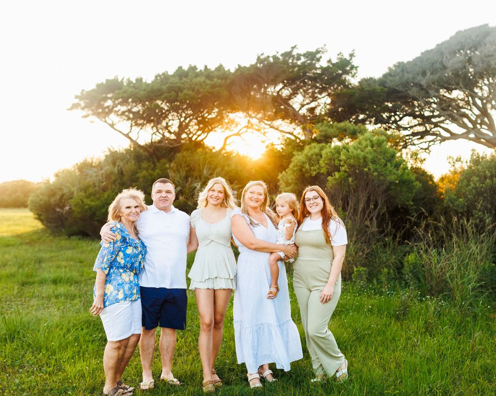 Multi-generational group standing together in a grassy field at sunset with trees in background.