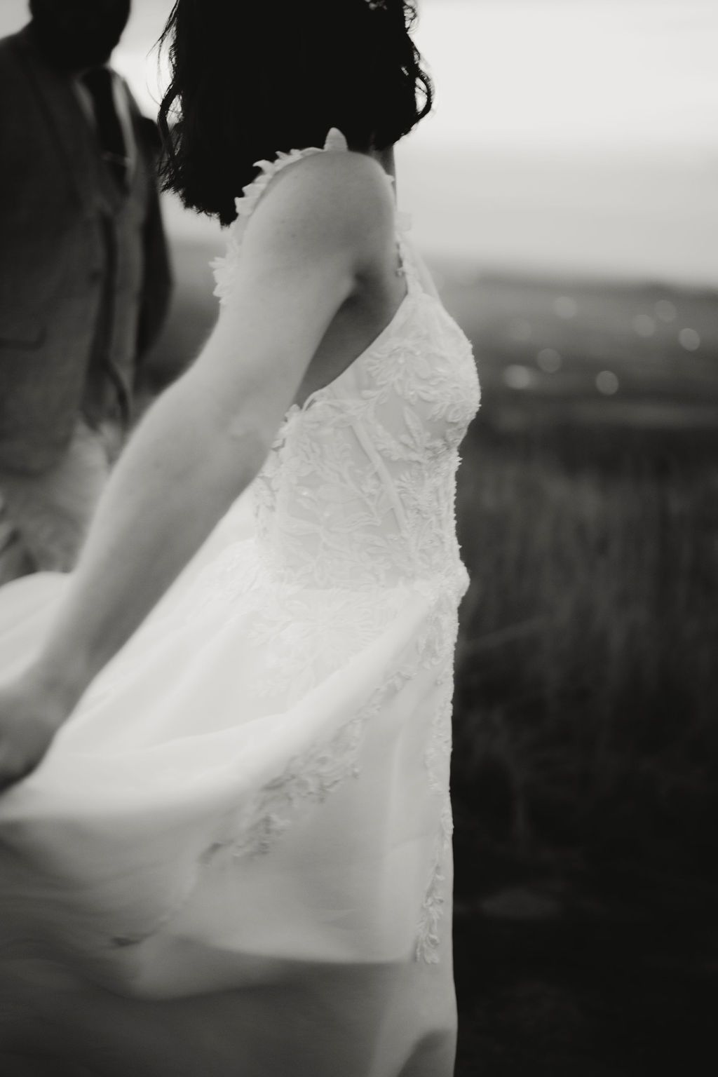A black and white photo shows a delicate white lace wedding dress flowing in a gentle breeze against a blurred landscape. Intimate weddings