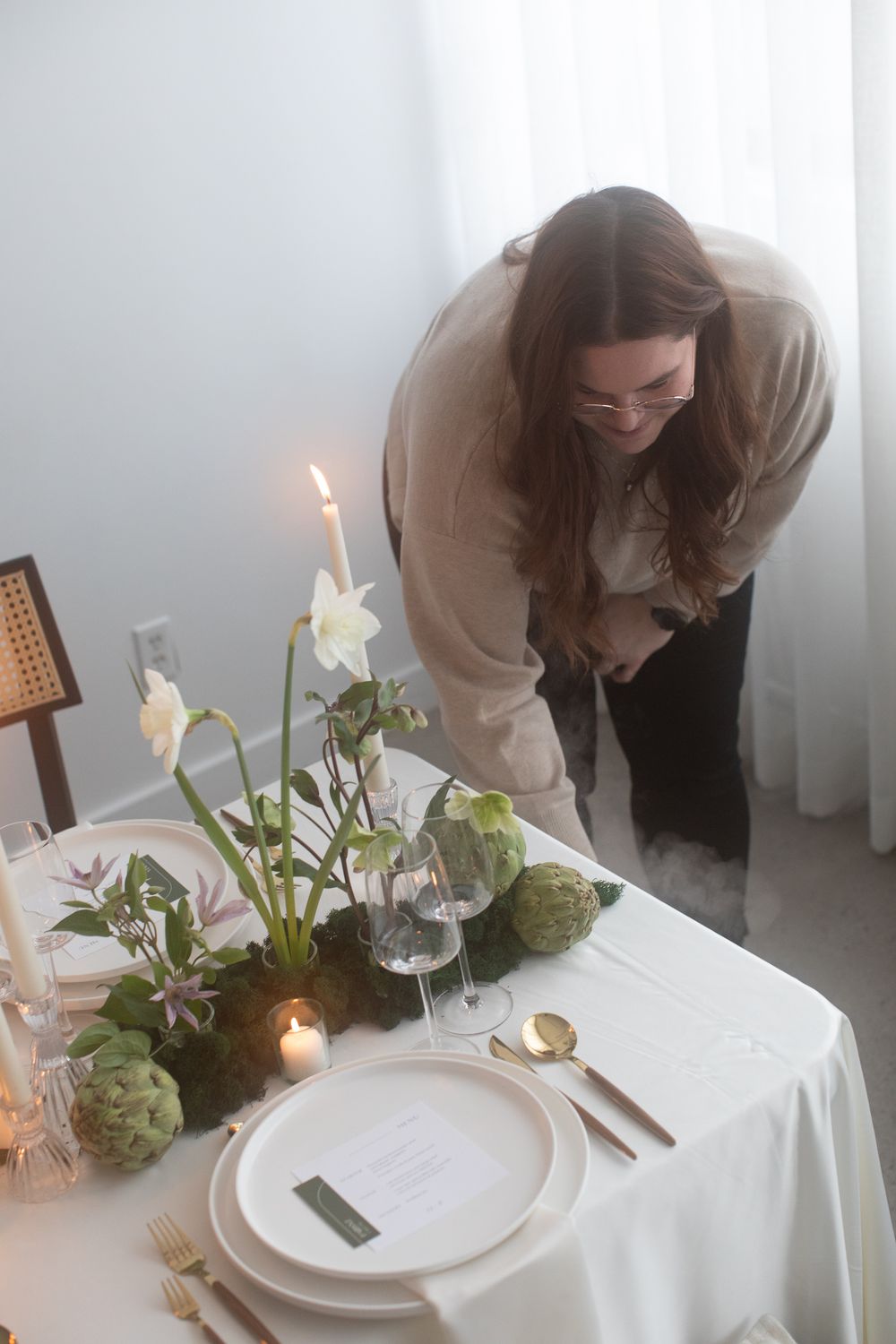 Woman in beige sweater arranging elegant green botanical wedding table setting with candles.