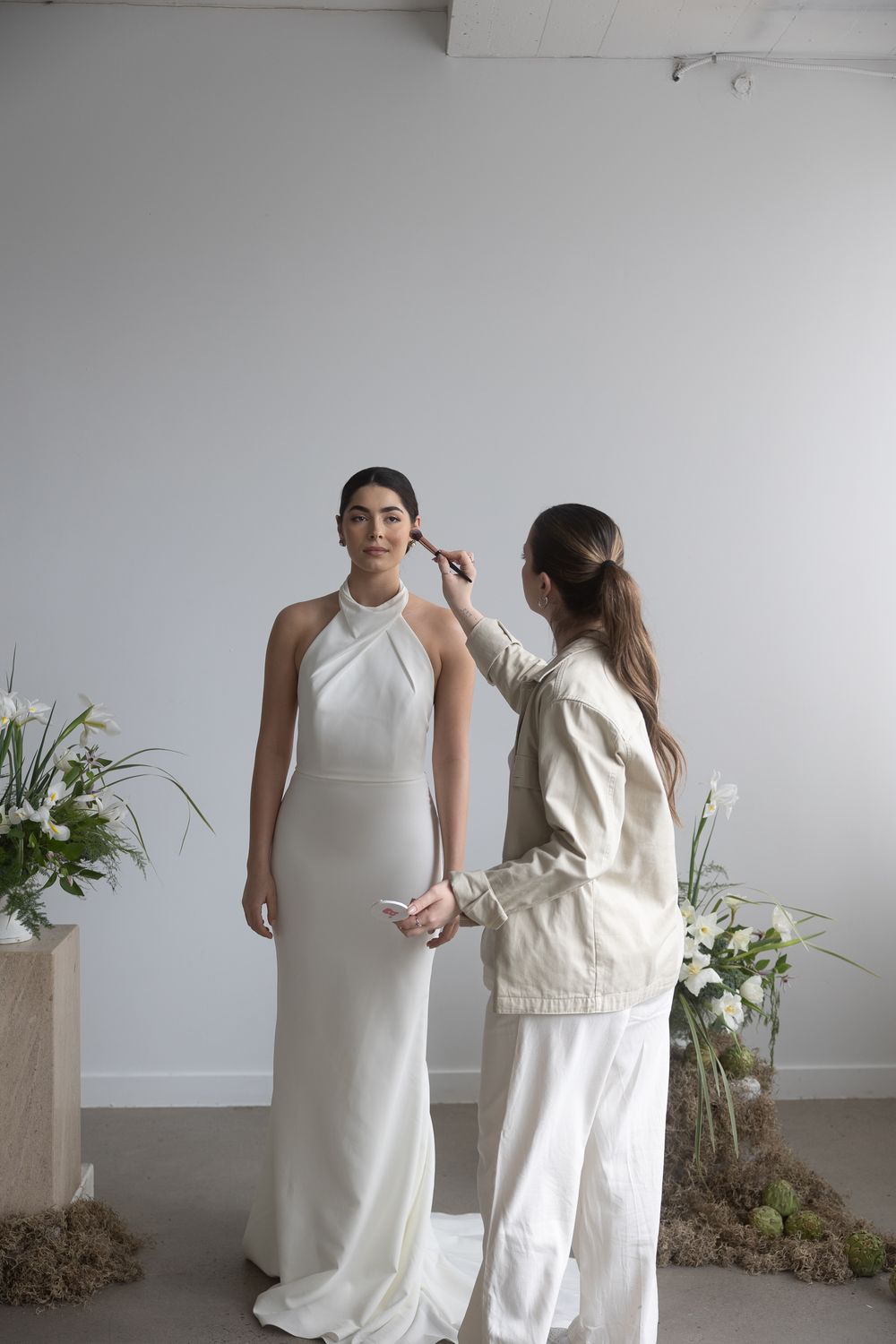 Bridal stylist adjusting hair of model wearing elegant white halter neck wedding gown.