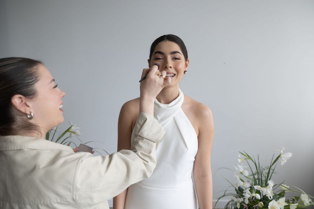 Makeup artist applying lipstick to bride in white halter wedding dress in studio.