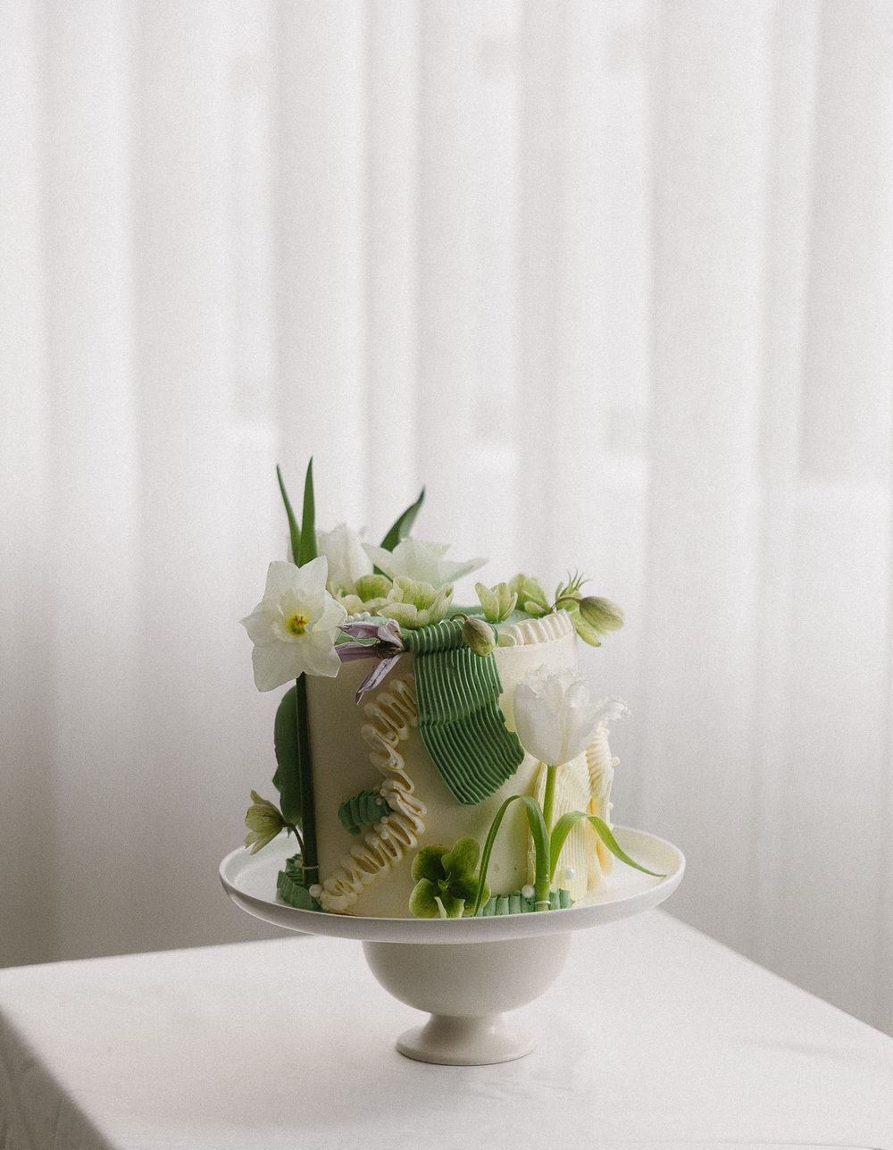 Elegant white wedding cake decorated with fresh green botanical flowers on white cake stand.
