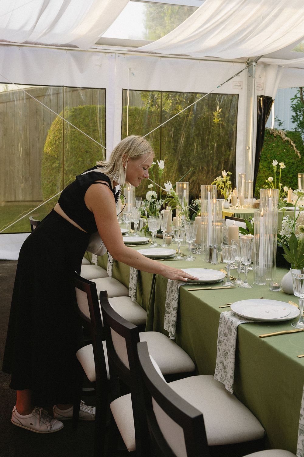 Wedding planner in black dress arranging a green and white tablescape inside an elegant tent venue.