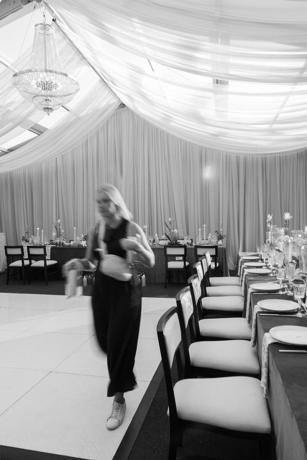 Black and white photo of a wedding planner moving through a draped tent reception venue with set tables.