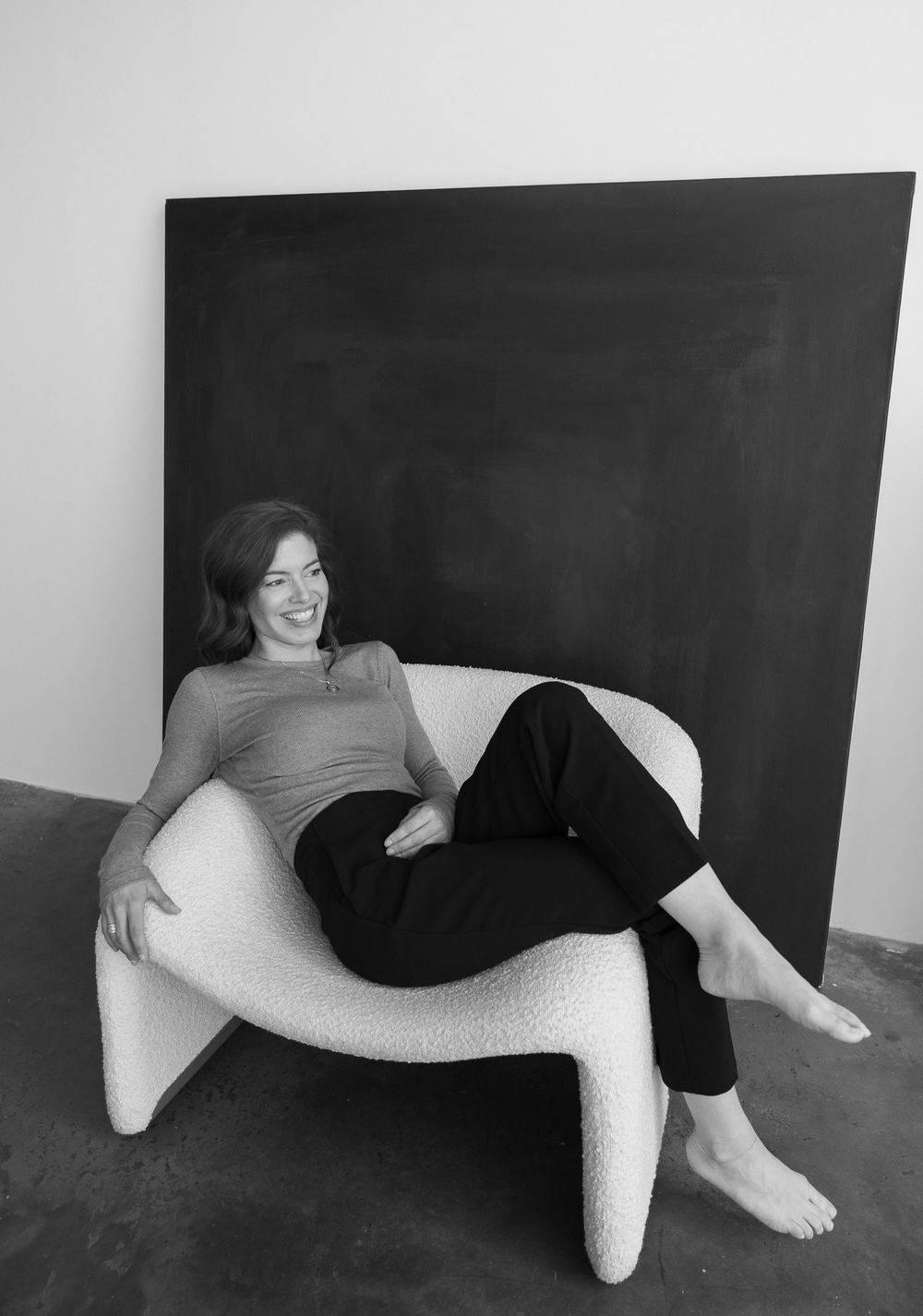 Woman relaxing in a modern white boucle chair, smiling, with a dark canvas artwork in the background.